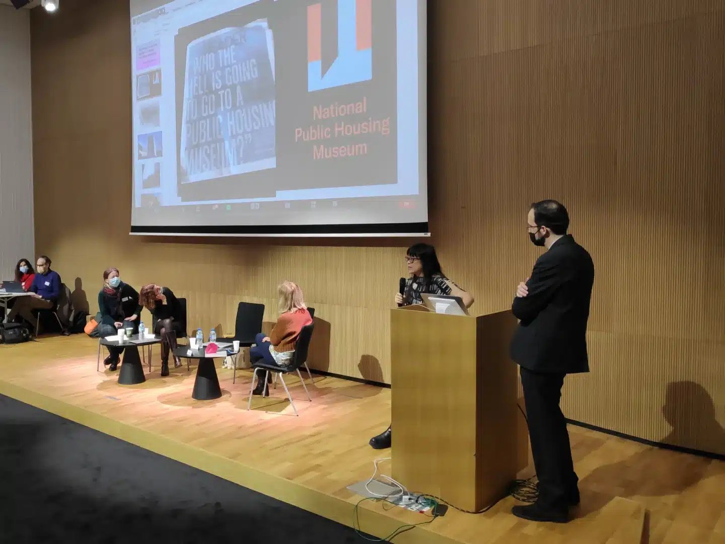 Intervenante à un colloque s'exprimant avec un micro devant sa présentation et plusieurs participants d'une table ronde