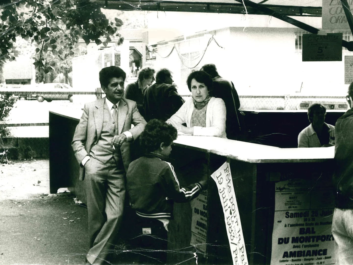 Photographie en noir et blanc. Un couple debout accoudé de part et d'autre d'un stand de nourriture d'une fête de quartier. Un enfant, de dos, passe devant eux.